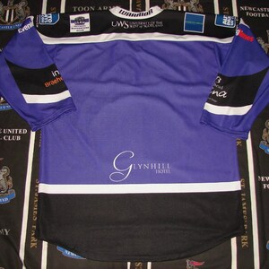 May include: A purple and black long-sleeved jersey with white accents. The jersey has the text "WARRIOR" and "UWS UNIVERSITY OF THE WEST OF SCOTLAND" on the front. The jersey also has the text "GLYNHILL HOTEL" on the front.