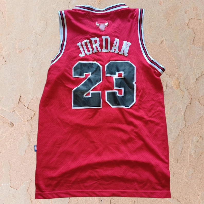 Puede incluir: Camiseta de baloncesto roja con ribetes negros y blancos. La parte posterior de la camiseta tiene el nombre "JORDAN" y el n&uacute;mero "23" en negro.