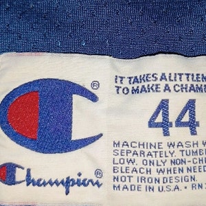 Puede incluir: Etiqueta Champion blanca con un logotipo azul y rojo, el texto "IT TAKES ALITTLEMORE TO MAKE A CHAMPION", el n&uacute;mero "44" e instrucciones de cuidado. La etiqueta est&aacute; sobre un fondo azul.