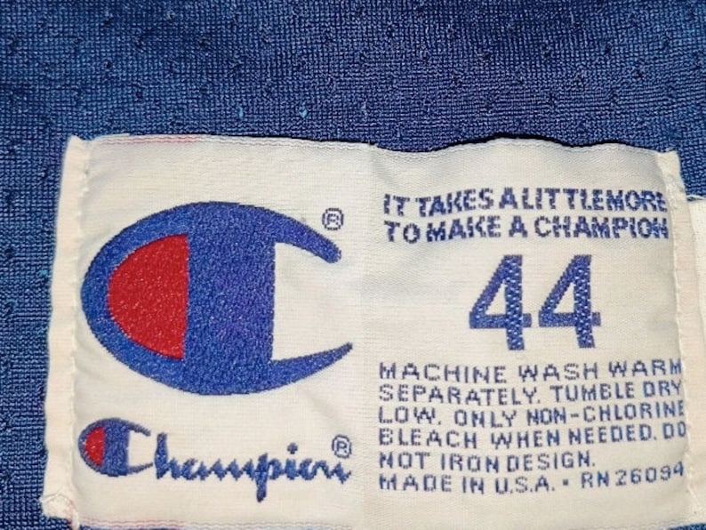 Puede incluir: Etiqueta Champion blanca con un logotipo azul y rojo, el texto "IT TAKES ALITTLEMORE TO MAKE A CHAMPION", el n&uacute;mero "44" e instrucciones de cuidado. La etiqueta est&aacute; sobre un fondo azul.