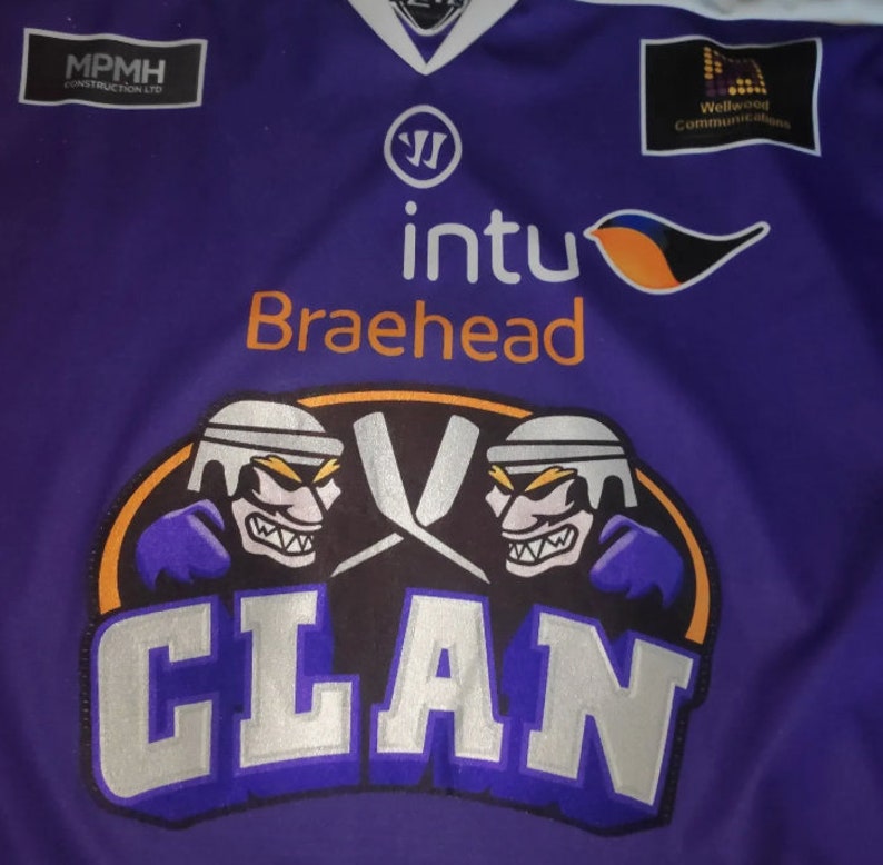 May include: Purple hockey jersey with the word "CLAN" in silver lettering on the front. The jersey has a logo with two hockey players and crossed hockey sticks. The jersey also has the names of sponsors, including "Braehead" and "Wellwood Communications."