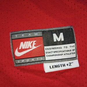 Puede incluir: Una camiseta roja Nike Team Sports con un logotipo blanco de Nike y el texto "TEAM NIKE SPORTS" y "M" en la etiqueta. La etiqueta tambi&eacute;n dice "ENGINEERED TO THE EXACT SPECIFICATIONS OF CHAMPIONSHIP ATHLETES" y "LENGTH +2".