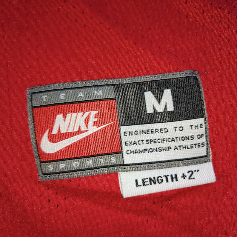 Puede incluir: Una camiseta roja Nike Team Sports con un logotipo blanco de Nike y el texto "TEAM NIKE SPORTS" y "M" en la etiqueta. La etiqueta tambi&eacute;n dice "ENGINEERED TO THE EXACT SPECIFICATIONS OF CHAMPIONSHIP ATHLETES" y "LENGTH +2".