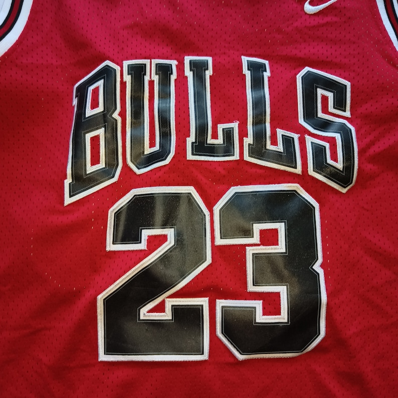Puede incluir: Camiseta de baloncesto roja con letras negras y blancas que dicen "BULLS" y el n&uacute;mero "23" en la parte delantera.