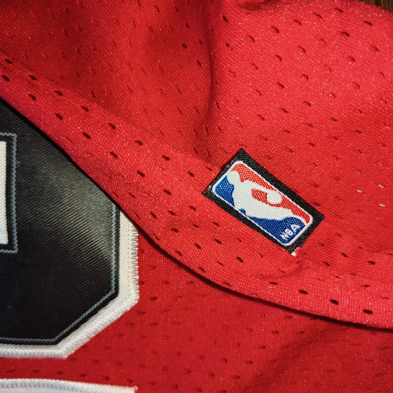 Puede incluir: Camiseta de baloncesto roja con un n&uacute;mero negro y un parche de logotipo de la NBA negro y azul.
