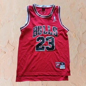 Puede incluir: Camiseta de baloncesto roja con ribetes negros y blancos. La camiseta tiene "BULLS" y "23" impresos en la parte delantera en negro. El logotipo de Nike est&aacute; en el hombro derecho y una etiqueta de talla est&aacute; en la parte inferior derecha de la camiseta.