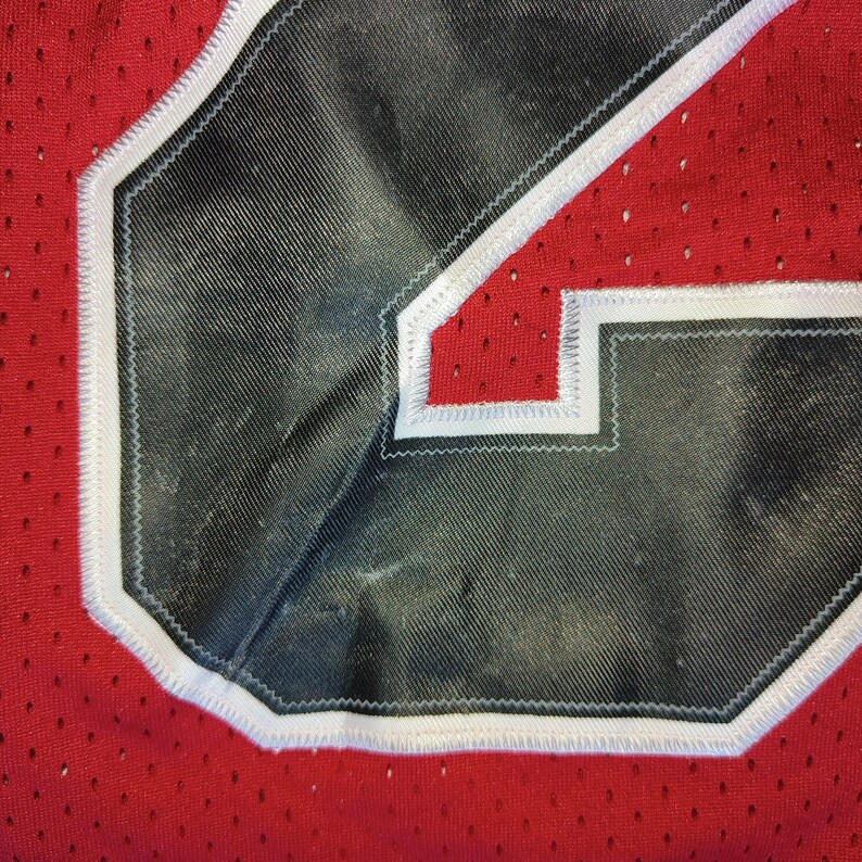 Puede incluir: Primer plano de un n&uacute;mero de camiseta negro y blanco con costuras blancas. El n&uacute;mero est&aacute; sobre un fondo rojo con un patr&oacute;n de malla.