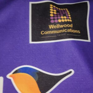 May include: Purple fabric with a black square logo that says "Wellwood Communications" and a yellow and purple geometric pattern. A black and orange bird with blue accents is also on the fabric.