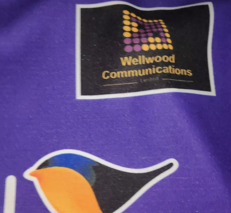 May include: Purple fabric with a black square logo that says "Wellwood Communications" and a yellow and purple geometric pattern. A black and orange bird with blue accents is also on the fabric.