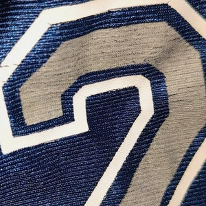 Puede incluir: Primer plano de una tela azul marino con un n&uacute;mero "2" blanco cosido sobre ella. El n&uacute;mero est&aacute; ligeramente descolorido y tiene un aspecto desgastado.