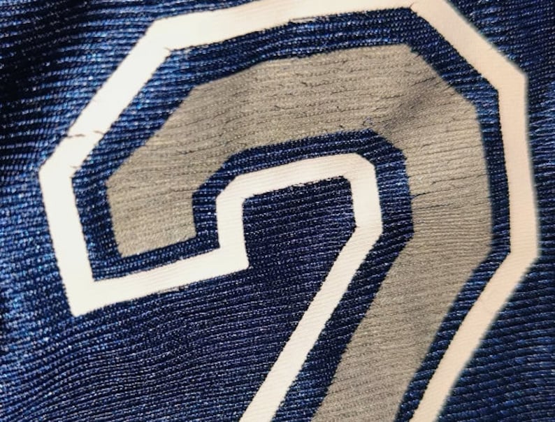 Puede incluir: Primer plano de una tela azul marino con un n&uacute;mero "2" blanco cosido sobre ella. El n&uacute;mero est&aacute; ligeramente descolorido y tiene un aspecto desgastado.