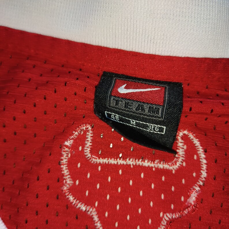 Puede incluir: Una camiseta de baloncesto roja con un logotipo de Nike blanco y la palabra "TEAM" en una etiqueta negra. La camiseta tiene un logotipo de cabeza de toro blanco en la espalda. La etiqueta tambi&eacute;n muestra la talla "M" y "3EG".