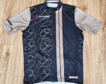 May include: Black and beige cycling jersey with a vertical stripe design. The jersey has a zipper closure and the text "STEINBACHER" on the front. The text "Durstig besser. Denk weiter." is also on the front of the jersey.