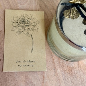 May include: A tan card with a black floral illustration and the text "Jess & Mark 07.19.2025". A decorative glass jar with gold accents is in the background. The card is on a wooden surface.