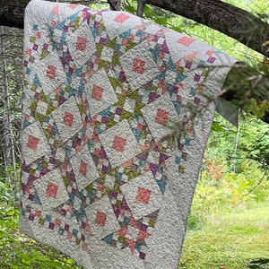Puede incluir: Una colcha blanca con un diseño de patchwork con cuadrados y estrellas de color rosa, verde, azul y morado. La colcha está colgada de una rama de árbol en un entorno boscoso.
