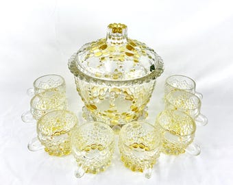 Può includere: Un set di ciotola da punch in vetro vintage con coperchio e otto tazze abbinate. Il set è trasparente con accenti gialli e un motivo strutturato a bolle. Le tazze hanno manici e piedini.