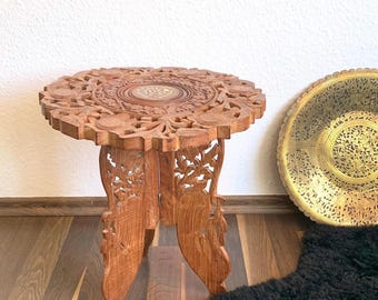 May include: A round, intricately carved wooden table with a floral design. The table has three legs and is made of dark brown wood. The table is sitting on a wooden floor with a black sheepskin rug in the foreground.