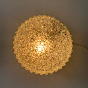 Pode incluir: Uma luminária de teto redonda de vidro com uma superfície texturizada. A lâmpada é composta por numerosos elementos de vidro transparente em forma de bolhas, criando uma forma esférica única. A luz é iluminada por dentro, emitindo um brilho quente.