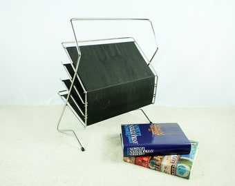 May include: A chrome magazine rack with black wire shelves. The rack is standing on a white carpet with two books stacked on the floor in front of it.