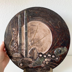 May include: A round ceramic wall plate with a brown, beige, and green glaze. The plate features a raised design of stylized plants and flowers.