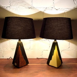 Puede incluir: Dos lámparas de mesa a juego con pantallas negras y bases geométricas. Las bases combinan tonos negros, dorados y marrones, creando un estilo art déco moderno. Las lámparas están colocadas sobre una superficie de madera contra una pared de ladrillo blanco.