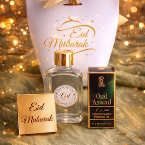 Può includere: Un set regalo per Eid Mubarak, con un flacone di profumo, una scatola di olio profumato concentrato e un sacchetto regalo. La borsa e una piccola carta quadrata mostrano le parole "Eid Mubarak" in oro. Il flacone di profumo ha un tappo dorato.