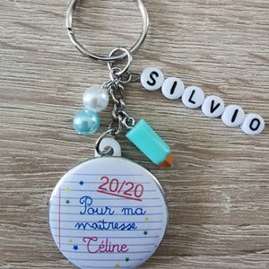 Peut inclure: Un porte-clés argenté avec un charme crayon bleu et blanc, un charme perle blanche et un charme blanc avec le mot "SILVIO" écrit en lettres noires. Le porte-clés a également un bouton rond blanc avec un "20/20" noir et rouge écrit dessus et le texte "Pour ma maitresse Celine" écrit en noir sur fond blanc.