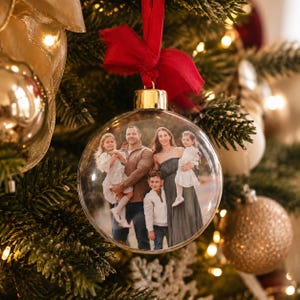 Op de afbeelding: Een doorzichtige, bolvormige glazen kerstbal met een afgedrukte foto van een gezin van vier personen. De kerstbal is voorzien van een goudkleurige metalen dop en een felrode stoffen strik en hangt aan een groene dennentak.