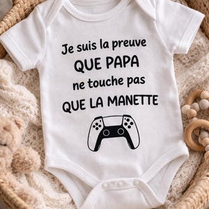 Peut inclure: Un body pour bébé blanc à manches courtes avec le texte noir "Je suis la preuve QUE PAPA ne touche pas QUE LA MANETTE" imprimé au-dessus d'une illustration graphique d'une manette de jeu vidéo. Le vêtement possède un col rond et des boutons-pression.