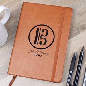 Puede incluir: Un diario de cuero marrón con un símbolo de clave de sol negro y el texto "John F. Murray VIOLA" en la portada.