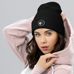 Peut inclure: Un bonnet noir avec un motif brodé blanc de deux masques de théâtre à l'intérieur d'un cercle.