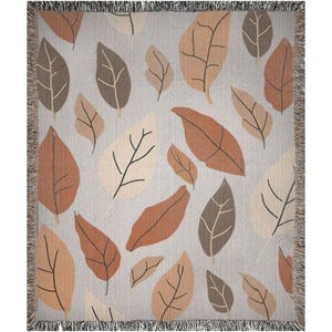 Puede incluir: Una manta tejida con un patrón de hojas marrones, beige y naranja sobre un fondo gris claro. La manta tiene flecos en todos los lados.