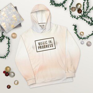 May include: A white hoodie with a faded orange and pink ombre design. The hoodie has a large pocket on the front and a drawstring hood. The text "MUSIC IN PROGRESS" is printed on the front of the hoodie in a brown rectangle with a musical note symbol.