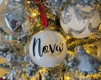 May include: A clear glass Christmas bauble with white feathers and the word "Nova" written in black calligraphy. The bauble is hanging on a Christmas tree with other decorations.