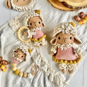 Puede incluir: Juguetes y accesorios de cordero de ganchillo en crema, rosa y amarillo. El conjunto incluye un sonajero con un anillo de madera, un clip para chupete con cuentas de madera y dos muñecos de cordero. Cada cordero lleva un vestido rosa.