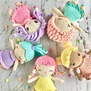 Puede incluir: Cinco muñecas de crochet con vestidos y cabello de diferentes colores. Las muñecas sonríen todas y tienen una expresión dulce. Están dispuestas en círculo sobre un fondo de madera blanco.