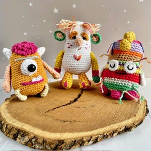 May include: Three colorful, handmade crochet monster dolls. The dolls are in shades of yellow, white, red, and green. Each doll has unique features, including large eyes, and whimsical details. They are displayed on a wooden slice.