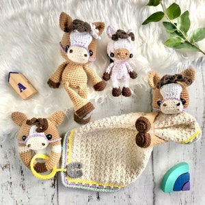 Puede incluir: Un conjunto de tres juguetes de caballo de crochet, que incluyen un caballo marrón y blanco con un anillo amarillo, un caballo marrón y blanco con una crin morada y un caballo rosa y blanco con una crin morada. Los juguetes están sobre una superficie blanca y peluda con un bloque de madera y un juguete de madera de colores arcoíris.