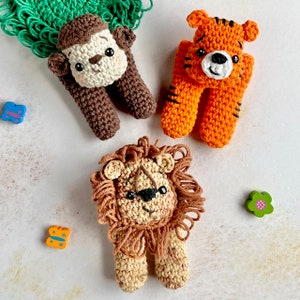 May include: Three crocheted stuffed animals: a brown monkey, an orange tiger, and a brown lion with a beige mane. The animals are all standing on a white surface.