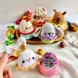 Op de afbeelding: Een verzameling gehaakte paaseieren dieren, waaronder een konijn, kuiken, lam en lama. De dieren zitten op een houten oppervlak met chocolade eieren die eromheen liggen. Het konijn houdt een chocolade ei in zijn pootjes.