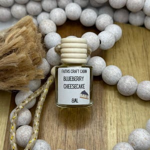 Peut inclure: Petite bouteille en verre avec un bouchon en bois et une étiquette indiquant "Faiths Craft Cabin Blueberry Cheesecake 8ml". La bouteille est entourée de perles blanches et d'une touffe brune.