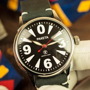 Peut inclure: Une montre-bracelet noire et blanche avec un boîtier argenté et un bracelet en cuir noir. Le cadran de la montre comporte de grands chiffres blancs et une aiguille des secondes rouge. Le cadran de la montre comporte également le mot "PAKETA" et le texte "Сделано в СССР".