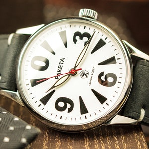 Peut inclure: Une montre-bracelet analogique blanche avec des chiffres et des aiguilles noirs. Le cadran de la montre porte l'inscription "PAKEta" et "Сделано в СССР". La montre est munie d'un bracelet en cuir noir.