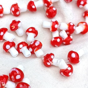 Può includere: Una collezione di perline di vetro a forma di fungo rosso e bianco. Le perline sono piccole e hanno un gambo bianco e un cappello rosso con punti bianchi.