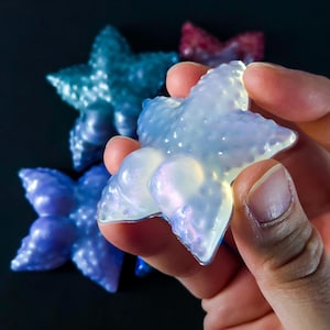 May include: A hand holds a translucent, iridescent starfish-shaped object. Other starfish in shades of blue and red are visible in the background. The objects have a textured surface and a pearlescent sheen.