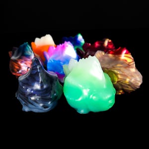 May include: A collection of colorful, abstract resin sculptures. The pieces feature a variety of colors, including blue, green, orange, and pink, with a glossy finish. The shapes are organic and irregular, with a textured surface. The background is black.