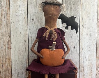 May include: A handmade Halloween witch doll with a brown pointed hat, purple dress, and an orange pumpkin. The doll has gray hair and is holding a black bat. It is sitting on a brown trunk and a wicker basket.