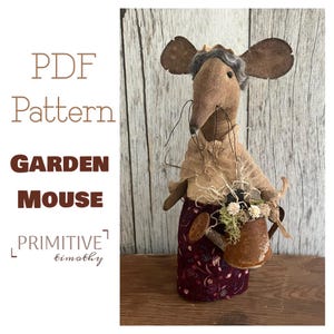 May include: A fabric mouse doll with large brown ears, gray hair, and long whiskers, wearing a tan top and a dark maroon floral skirt. The mouse holds a small, rusted metal watering can filled with dried flowers and moss. Text reads PDF Pattern GARDEN MOUSE PRIMITIVE timothy.