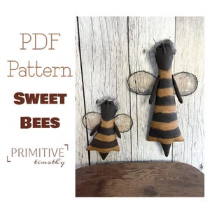 May include: Two fabric bee dolls with brown and tan horizontal stripes, gray hair, and thin wire antennae. The text reads: PDF Pattern SWEET BEES PRIMITIVE timothy. The dolls have dark brown faces and translucent, textured wings on their backs.