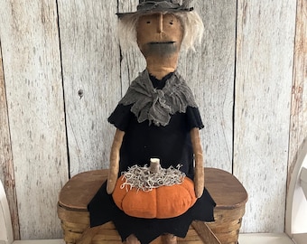 May include: A handmade Halloween witch doll with a black hat, dress, and boots. The doll is holding an orange pumpkin with a straw-like stem. The doll is sitting on a brown basket. The background is a light wood panel.